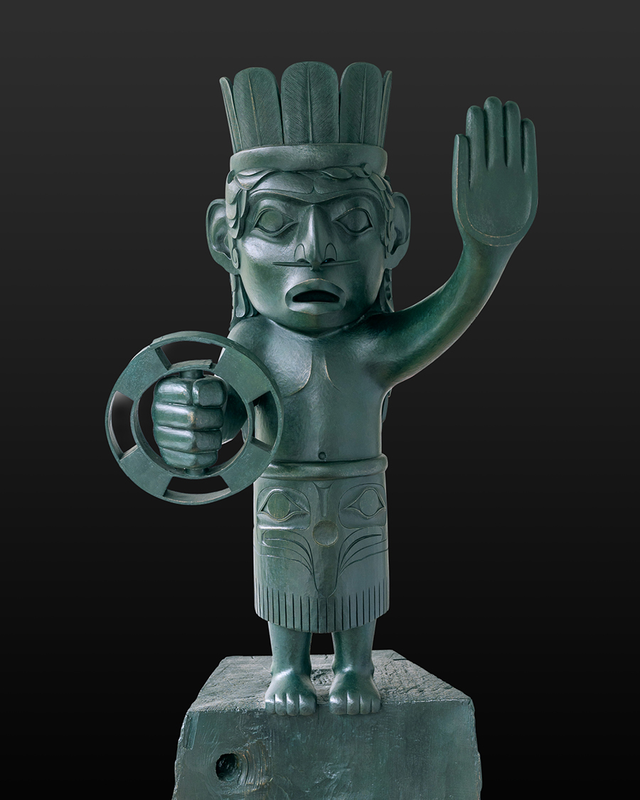 Green-painted carved figure of a human with feathered headdress, holding a circular object in one hand and raising the other in a gesture, by Jim Hart.