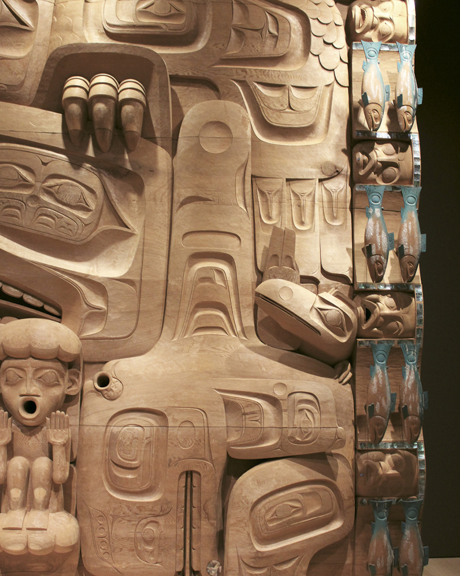 Close-up of the right side of Jim Hart’s carved cedar panel, showing intricate Haida designs, human and animal forms, and turquoise-painted salmon.