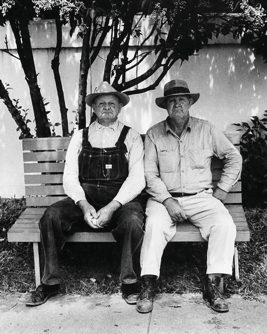 Two elderly men sit side by side on a wooden bench outdoors. One wears overalls and a plaid shirt, the other a button-down shirt and light trousers. Both wear wide-brimmed hats and have serious expressions. Trees and a white wall are in the background.