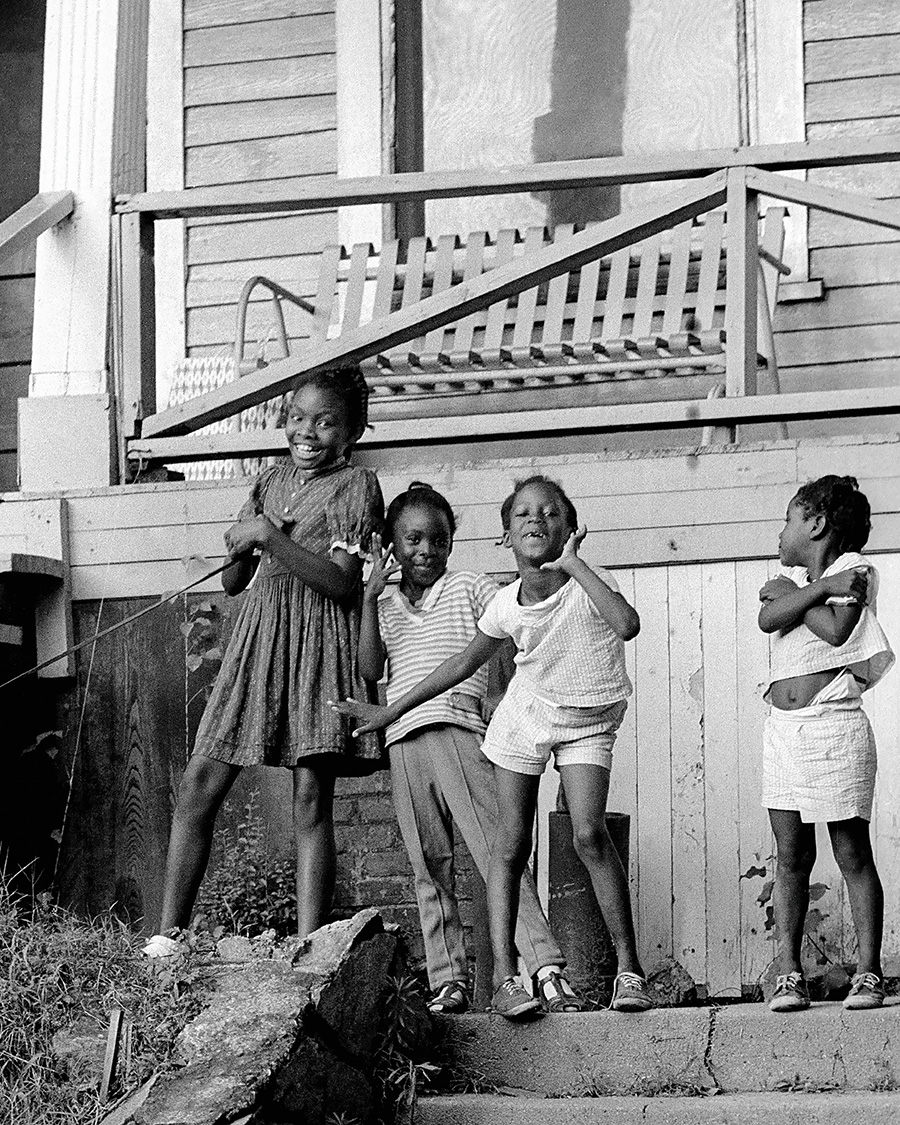 Four young girls stand on the steps of a wooden house, smiling and striking playful poses for the camera. They appear to be enjoying themselves on a sunny day, with a porch swing visible behind them.