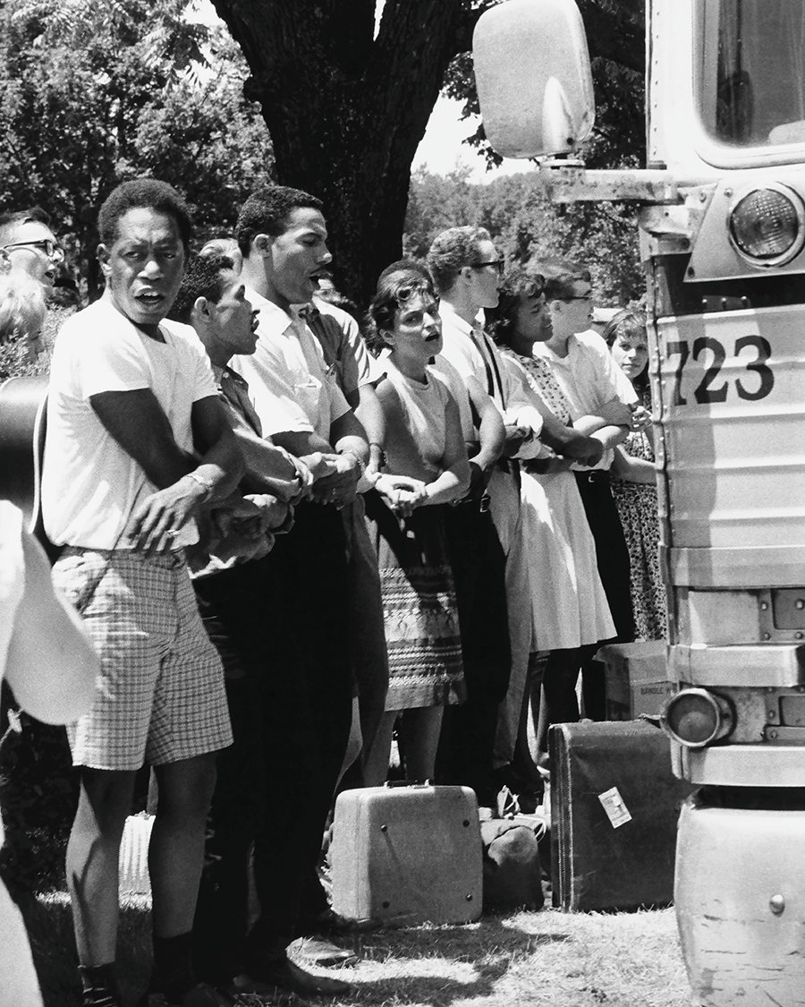 A racially diverse group of men and women stand arm-in-arm in front of a bus, participating in a civil rights protest. The group looks determined and unified, with suitcases placed on the ground nearby, suggesting travel or a march.