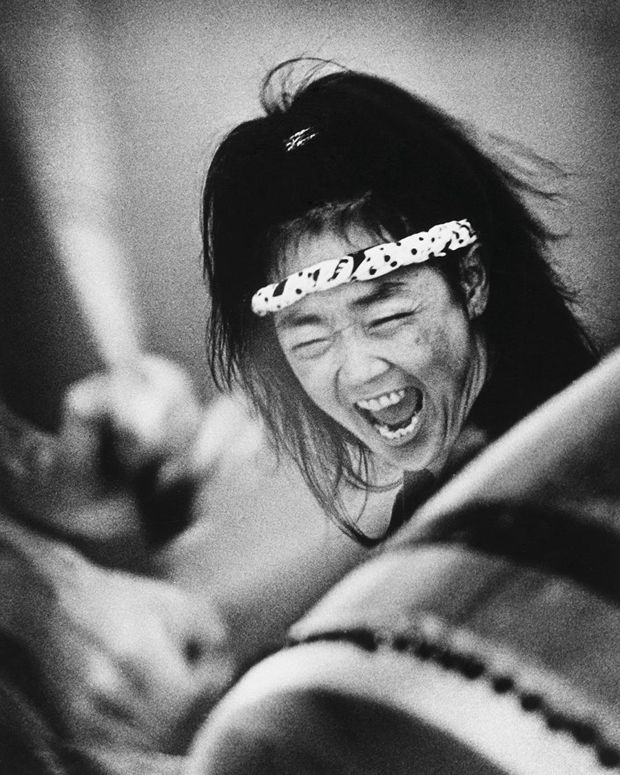 A woman with a headband and expressive face passionately plays a large traditional taiko drum. Her mouth is open as she shouts or sings, caught in an intense moment of performance. The image conveys motion and emotion.